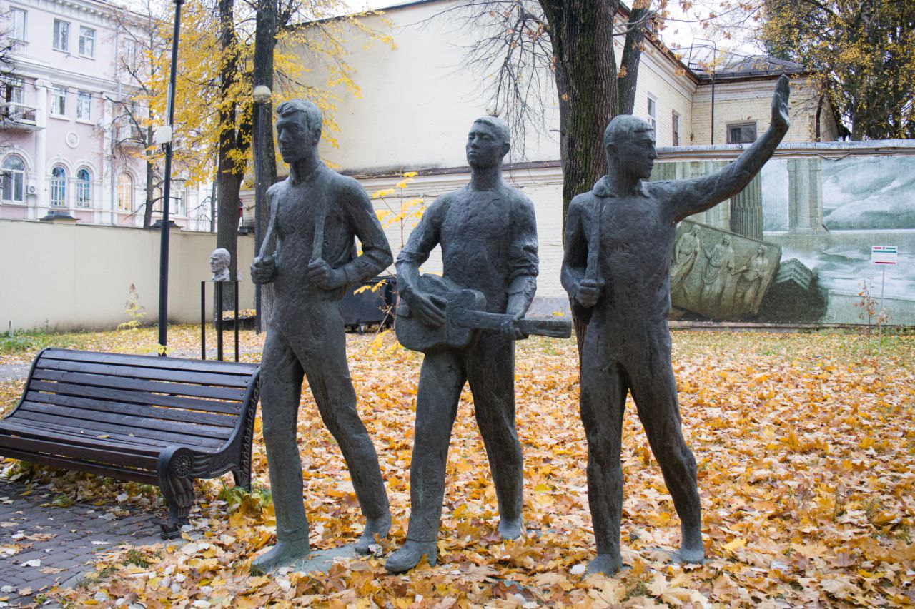 Призывники: На переднем плане скульптурная композиция. Три фигуры в полный рост размещены на едином основании неправильной формы. Молодой человек слева держит двумя руками лямки рюкзака. В центре композиции стоит молодой человек, выставив правую ногу вперед, в его руках гитара. Молодой человек справа держит лямку рюкзака. Он повернул голову влево, его левая рука поднята вверх, а левая нога отведена назад. Слева от многофигурной композиции скамейка, на заднем плане газон с деревьями и стены парка.